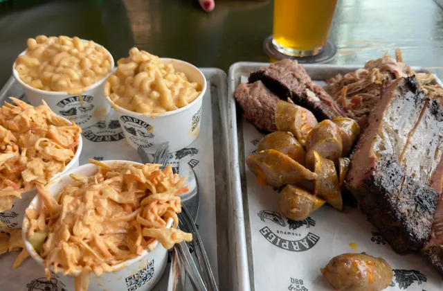 Close-up photograph of barbecue meats, sliced sausage, macaroni cheese and coleslaw on trays, with a cropped beer glass at Warpigs.